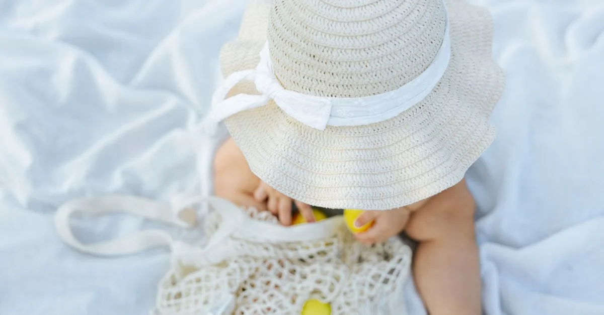 Bébé adorable en chapeau de paille explorant un sac en filet sur une douce couverture blanche.